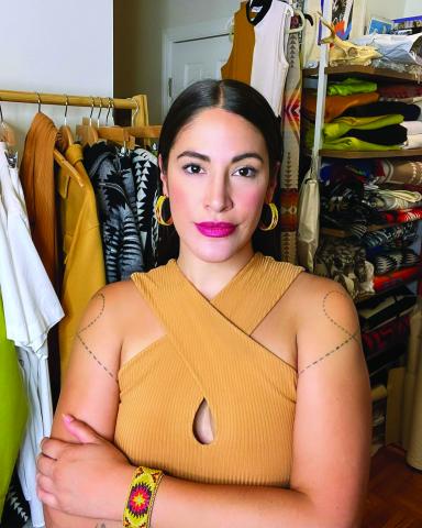 Woman with hair pulled back wearing a sleeveless yellow dress posing in front of a studio full of clothing.