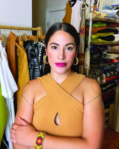 Woman with hair pulled back wearing a sleeveless yellow dress posing in front of a studio full of clothing. 