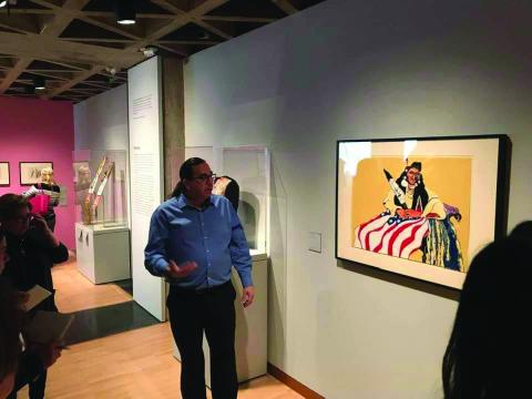 Man in blue shirt standing in front of painting in museum and gesturing while talking. 