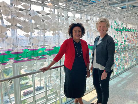 Woman with short curly brown hair in red jacket next to woman with short blonde hair in gray suit in front of glass sculpture.