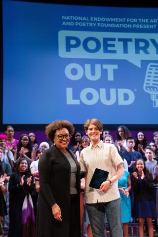 Two women, one middle aged and one teenaged, stand side by side smiling. The teenager holds an award