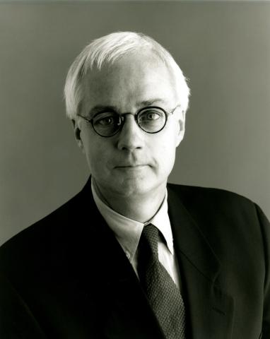 Portrait of white man with white hair wearing round black-framed glasses and wearing a dark suit. 