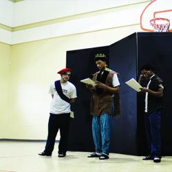 Three young men wearing costumes read from scripts in a gymnasium