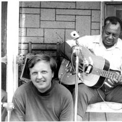 A man sits smiling on a porch step while a man plays guitar into a microphone while sitting behind him