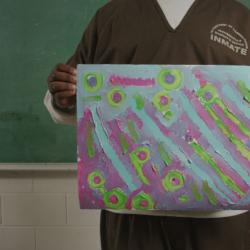 A young man in prison garb holds up a brightly colored painting that he made