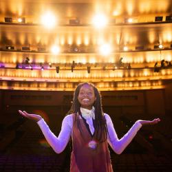 photo of young Black woman with long braids standing on stage with her back to audience and lights blazing behind her