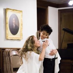 A smiling woman in white kneels next to a young boy in a tuxedo