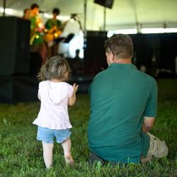 A toddler girl standing in the grass next to her sitting father as they watch an outdoor performance