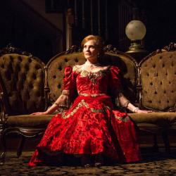 Marg Helgenberger in character as Regina Giddens on an early 1900s sofa wearing a red dress