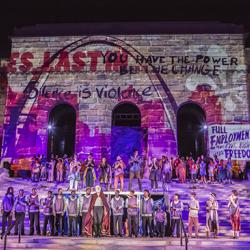 Performers line the steps of s stone building with art projections on it at night