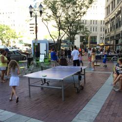 People on the sidewalk of a city doing hula hoop near a ping-pong table.