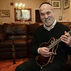 Andy Statman sits on a leather couch holding a mandolin in his hands. A clarinet can be seen standing on the floor behind him.