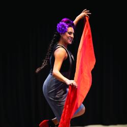 Woman with long braided hair and purple flower over her ear dancing on stage with a long orange scarf
