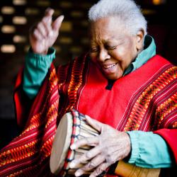 An older woman sits playing a talking drum. She is wearing brightly colored clothing and smiling as she gets ready to strike the drum again in song.