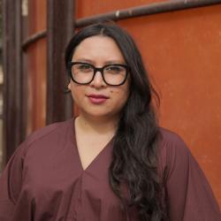 Portrait of woman with long dark hair wearing black-rimmed glasses and wearing a purple shirt. 