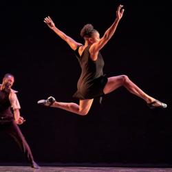 While a male-appearing dancer executes a move in the background, in the foreground a female-appearing dancer executes a leap.