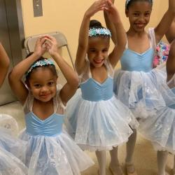 A group of young girls in leotards and tutus hold their hands above their heads in a ballet pose