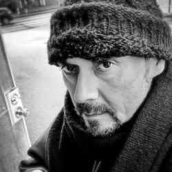 Black and white photo of man with a goatee wearing wool hat and a heavy coat standing in doorway. 