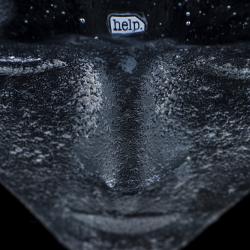 detail from a mask which is painted black and has a scrap with the word help embedded in the forehead of the mask