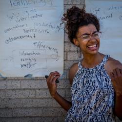 photo of Kendra Kreuger, who is a young Black woman with her hair pulled back and wearing glasses. She stands in front of large sheets of paper covered with text.