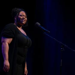 A young woman in a black dress speaks at a microphone