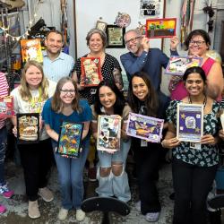 A group of teachers stand for a group photo, smiling and holding up their artwork.