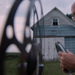 an older man with a long white beard stands outside a barn loading a reel onto an old fashioned film projector