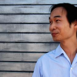 headshot of smiling Asian man in half-profile against background of weathered wood siding
