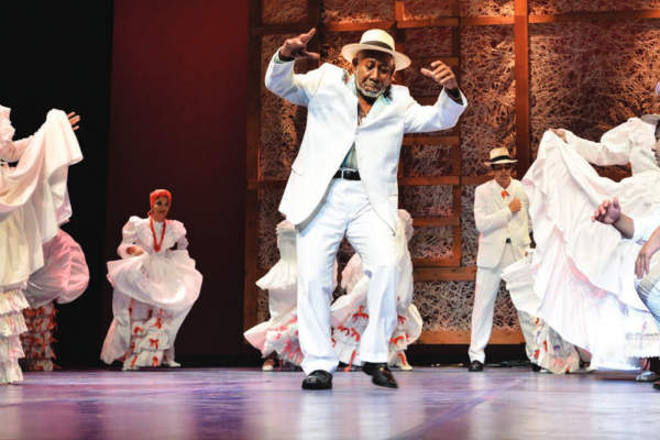 Men and women in white outfits on stage dancing to percussion from drummers at the edge of the stage