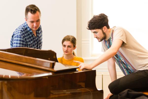 female student sits at piano with two men looking at sheet music with her