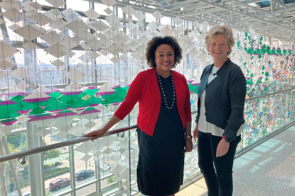 Woman with short curly brown hair in red jacket next to woman with short blonde hair in gray suit in front of glass sculpture.