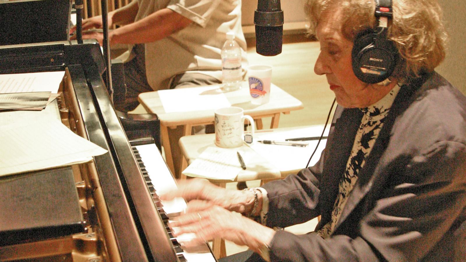 Marian McPartland Woman wearing headphones playing piano with African-American man behind her playing piano.