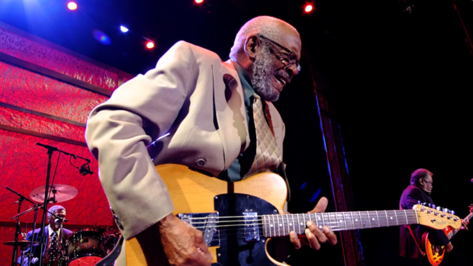 Man in a beige suit plays the electric guitar with a man on drums and another man on guitar in the background