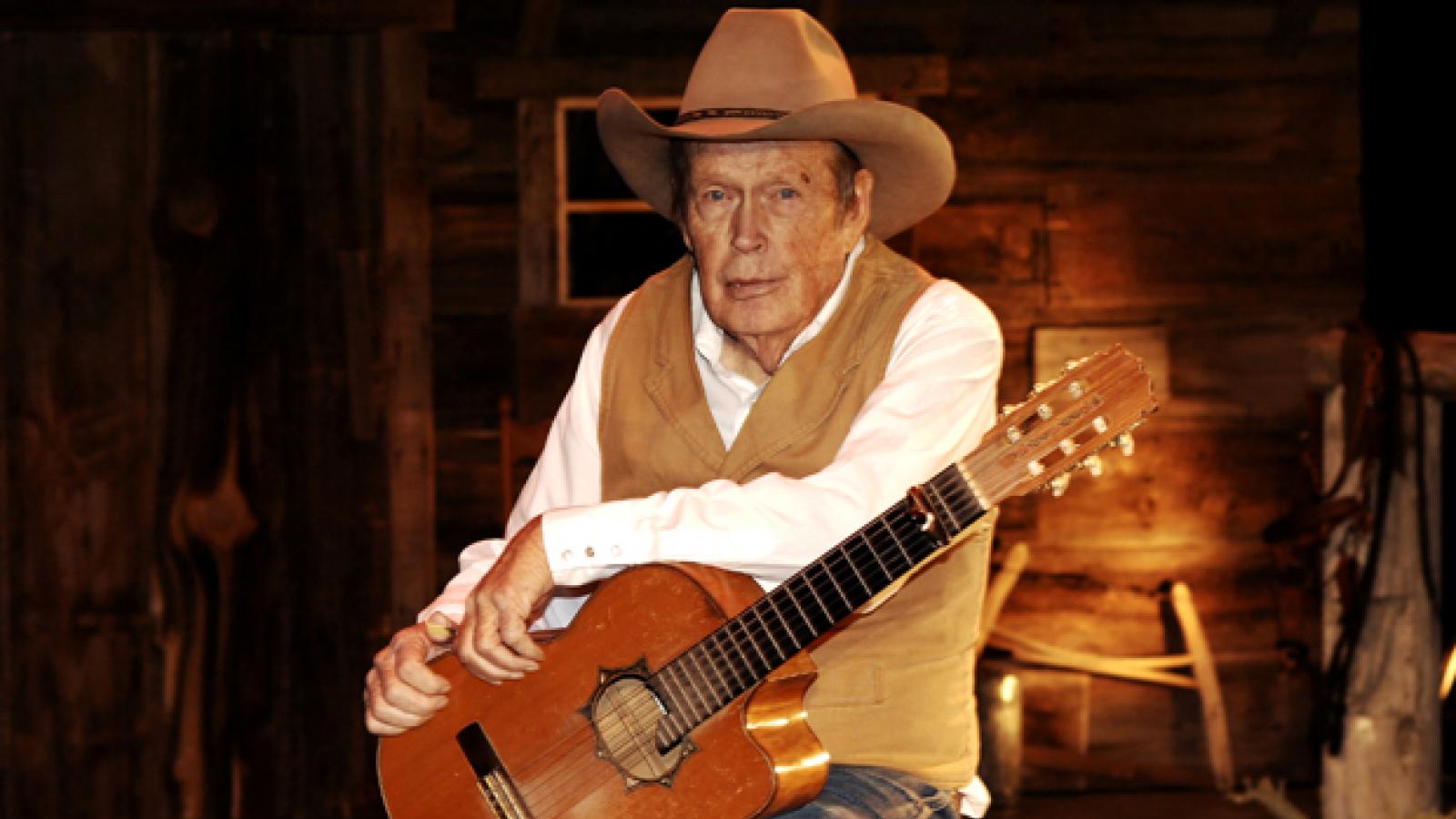 A man in a brown cowboy hat and best sits on a stool, holding a guitar.