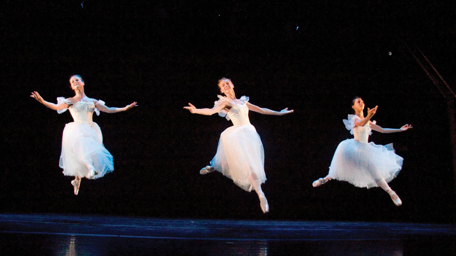 Three female ballerinas jumping onstage in white outfits as they perform. 