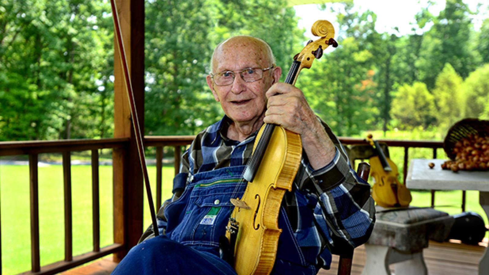 A man sits on a porch wearing overalls and holding a fiddle