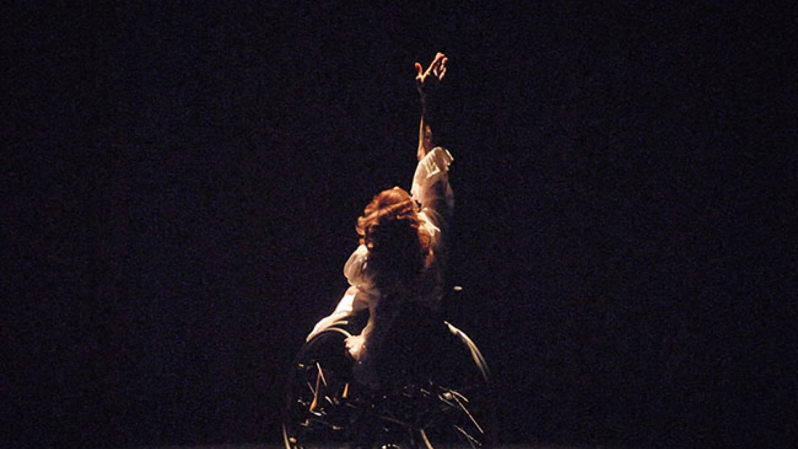 A dancer in a wheelchair lifts one hand upward as she performs onstage