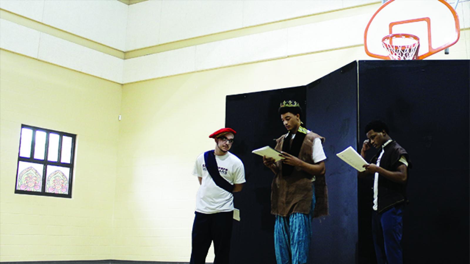 Shakespeare in American Communities Three young men wearing costumes read from scripts in a gymnasium