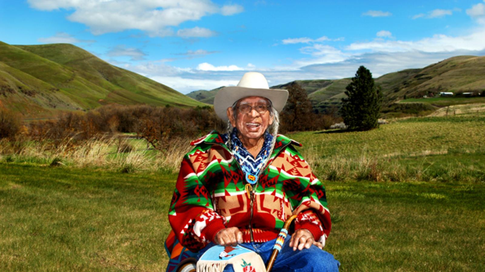 A man in a cowboy hat and colorful jacket sits in an open field.