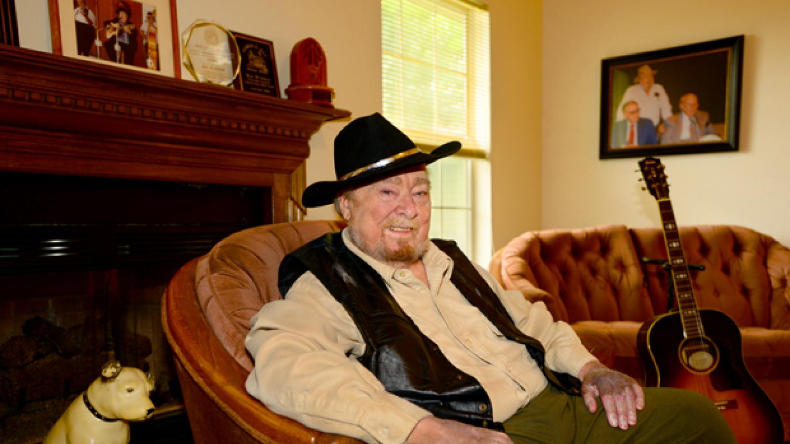 Mac Wiseman A man wearing a cowboy hat sits in a chair with a guitar next to him.