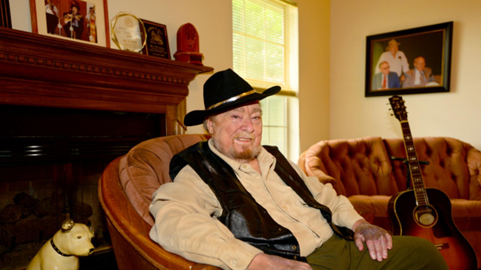 A man wearing a cowboy hat sits in a chair with a guitar next to him.