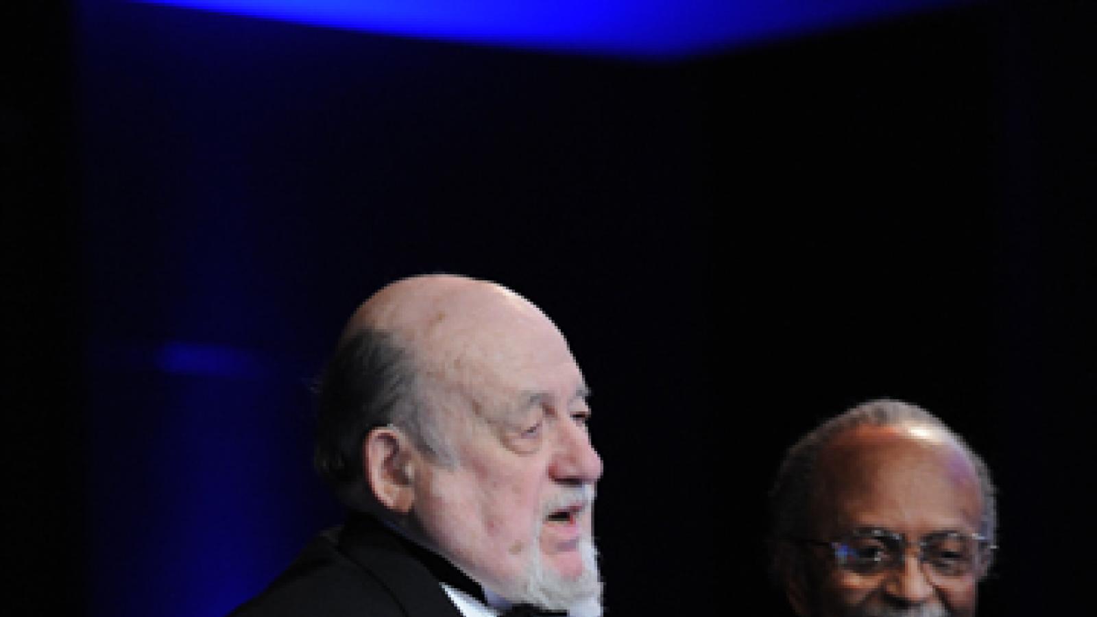 A man in a tuxedo speaks from a podium while another man in a brightly colored tie looks on smiling