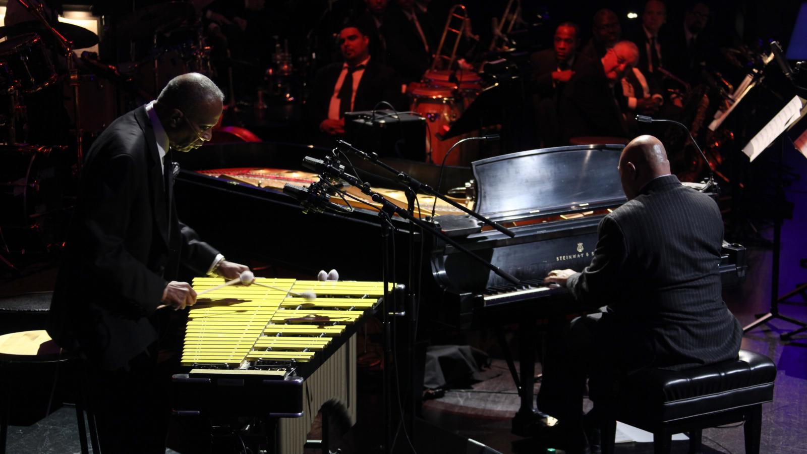 A man performs on the vibraphone next to another man performing on the piano.