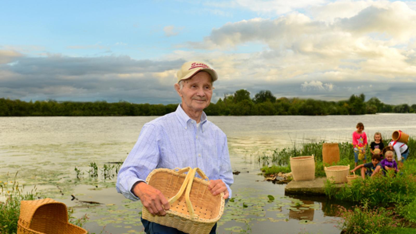 A man wearing a baseball hat holds a basket and stands in front of a lake, with several other baskets and children playing in the background.
