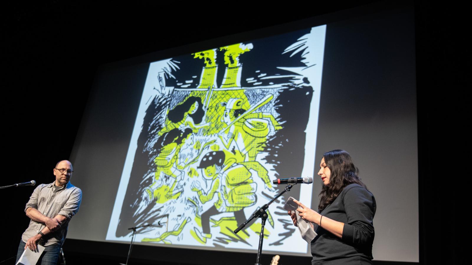 Daniel Kraus and Fernanda Melchor A man and a woman stand before microphones in front of a screen with a graphic novel image on it