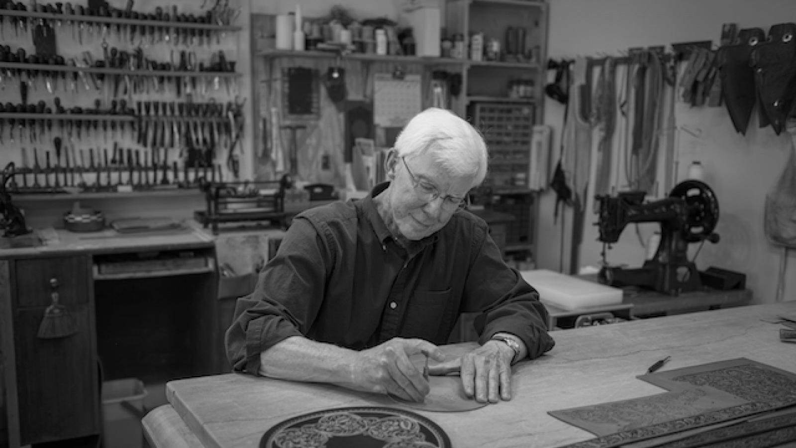 a gray-haired man sitting in a workshop working on a piece of leather