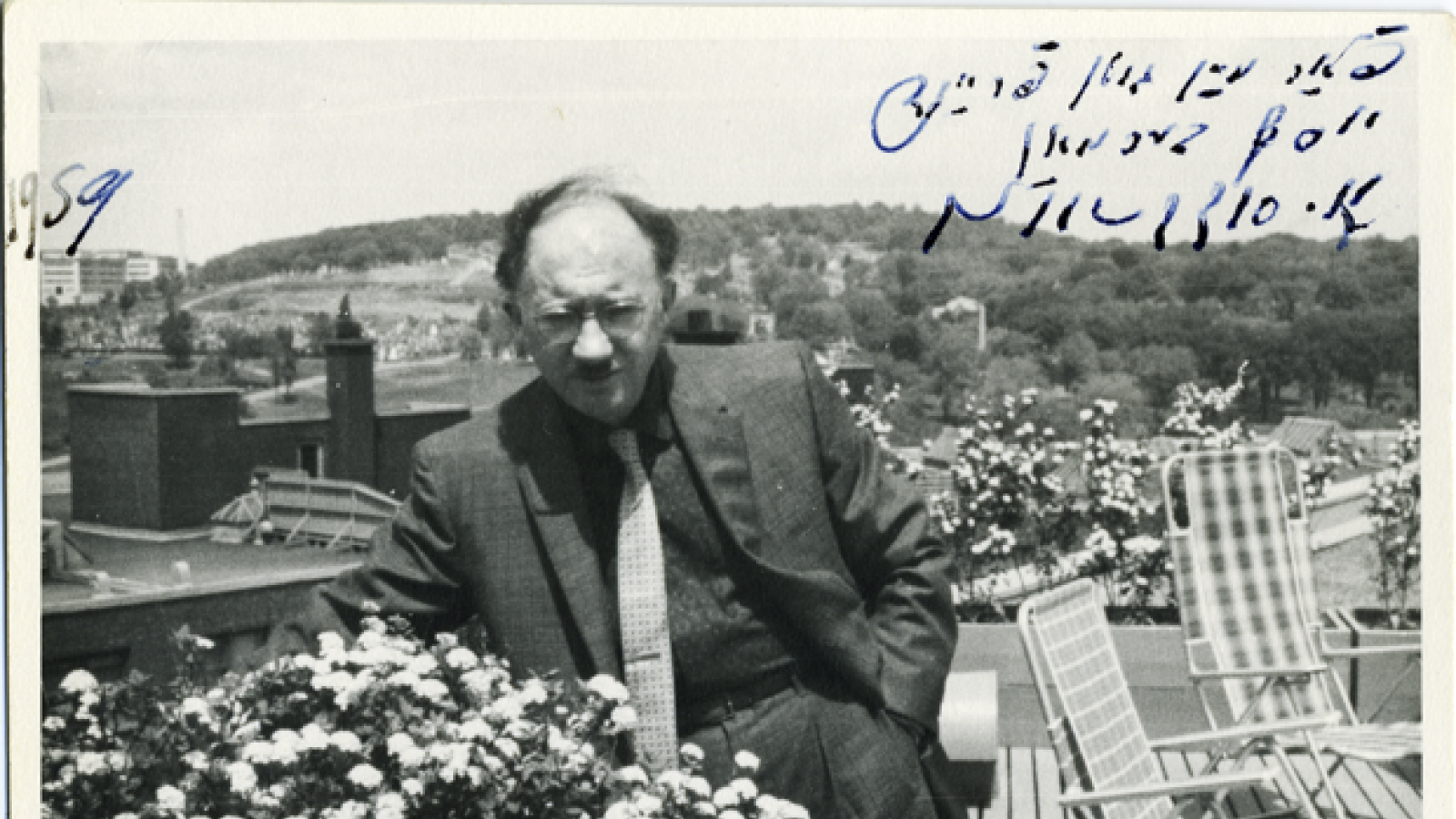 Black and white photo of a man wearing glasses and a suit standing on a balcony