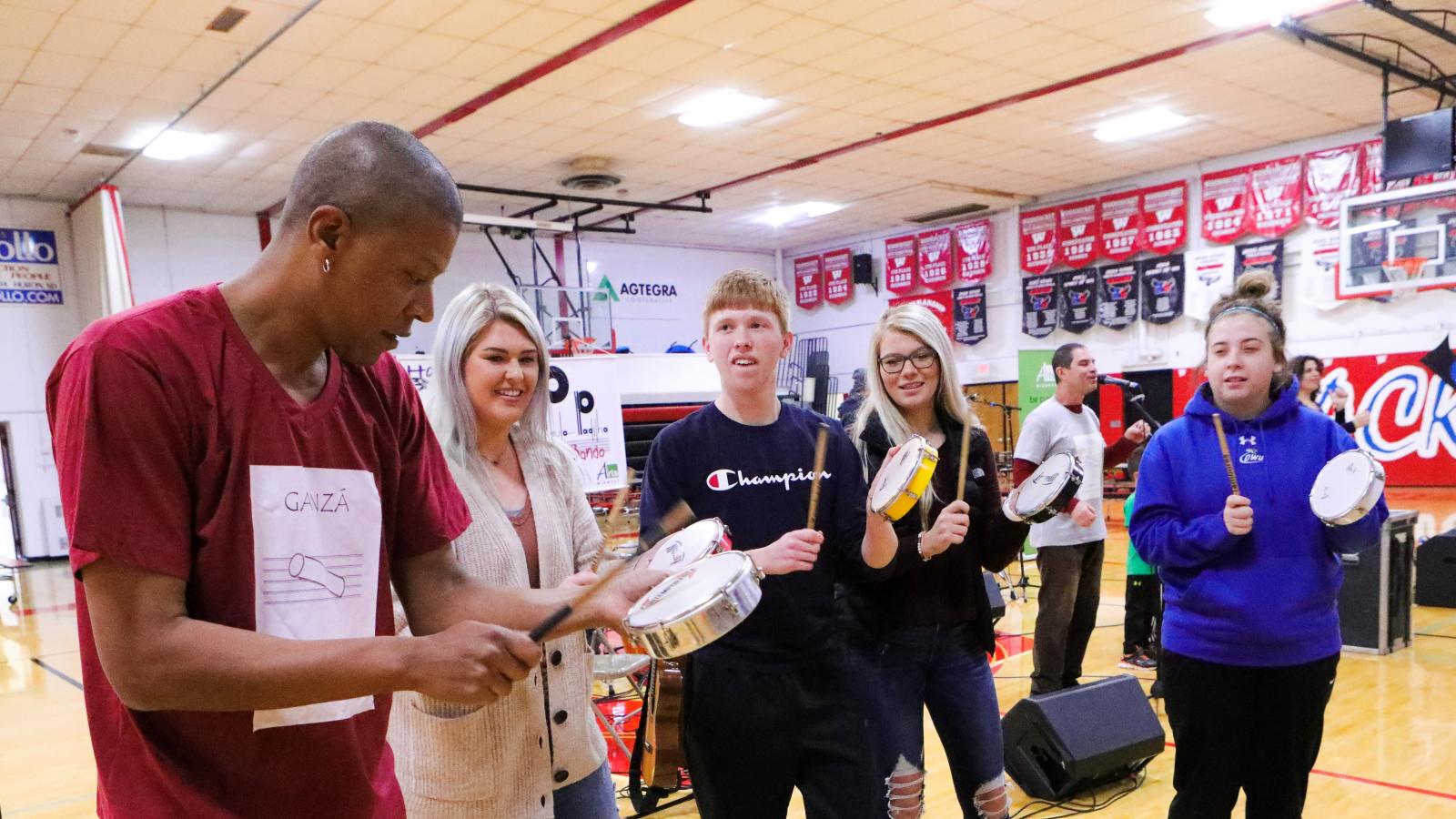 Samba Sam A man shows a group of teenagers in a high school gym how to play a Brazilian instrument