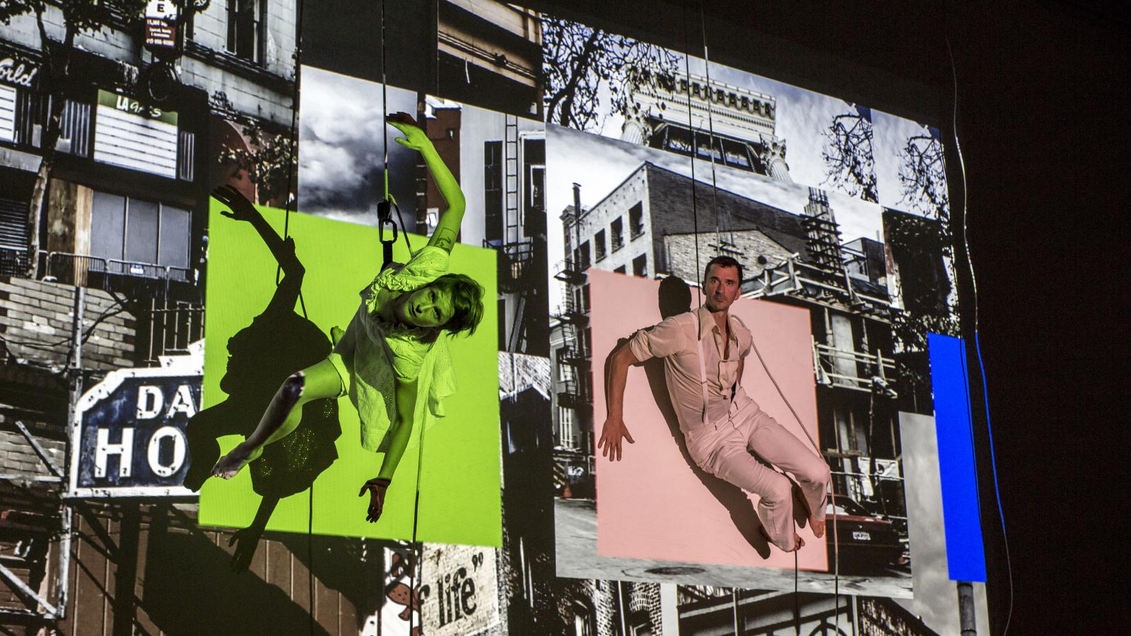 Two dancers suspended against a side of a building with video projections in the background