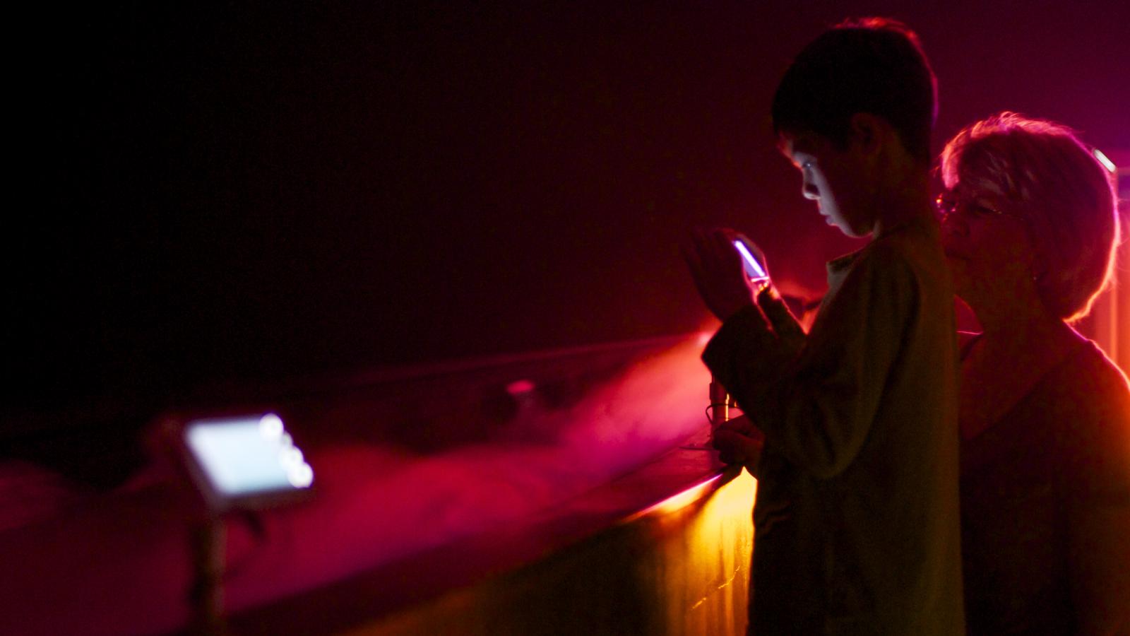 A boy holds a cellphone with an older woman behind him at a red cloud in front of him.